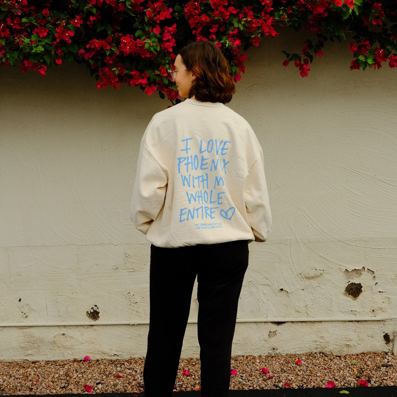 Person wearing a white sweatshirt with colorful text, standing in front of a wall with red flowers.