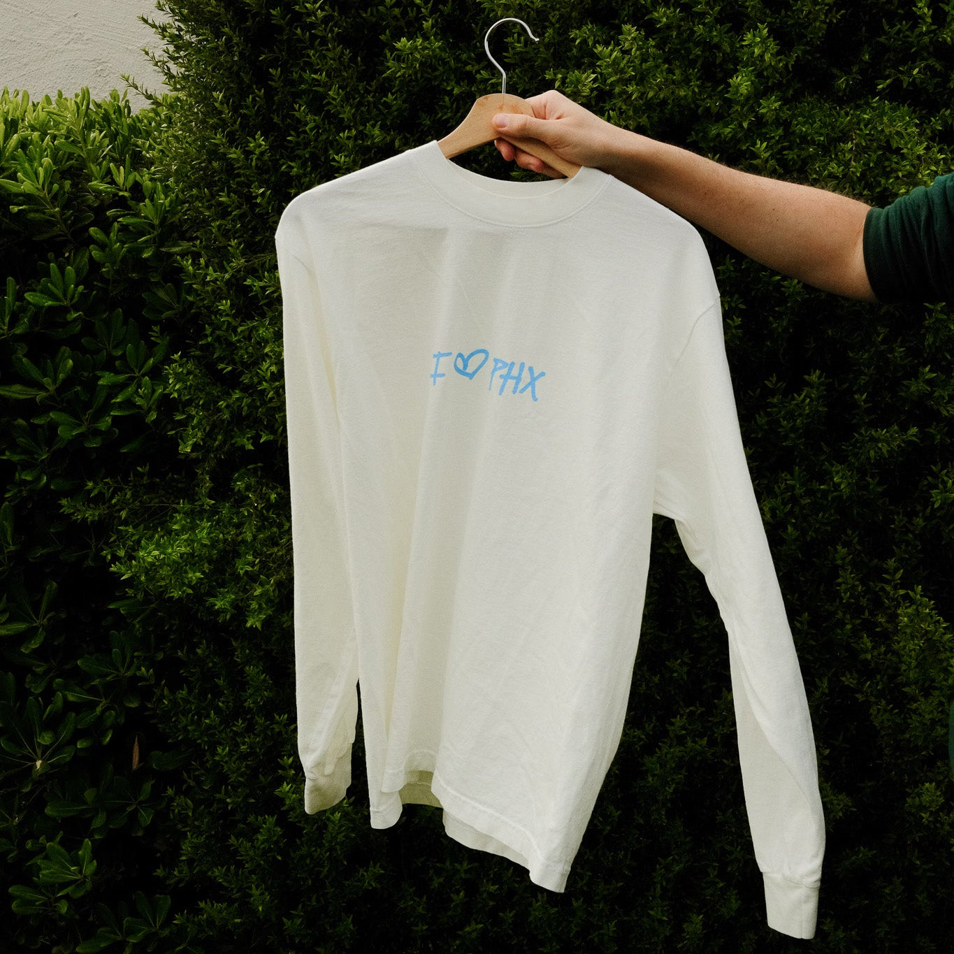 White sweatshirt with text on a hanger against a green bush background